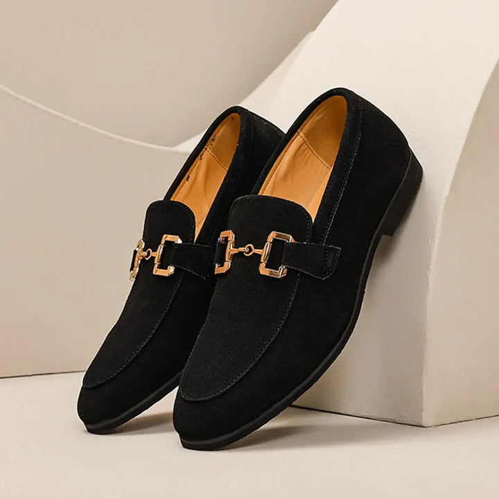 A pair of elegant black genuine leather loafers with gold horsebit detailing on the uppers, displayed on a cream-colored surface and background.