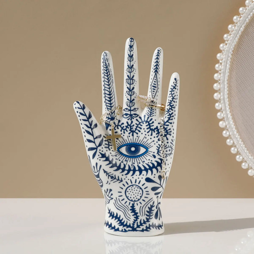 A ceramic jewelry stand in the shape of a hand, adorned with blue botanical and evil eye decor, holds gold rings. A round, pearl-trimmed mirror is partially visible on the right against a beige background.