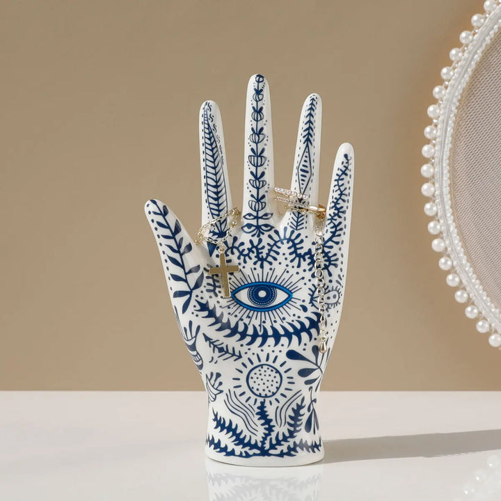 A ceramic jewelry stand in the shape of a hand, adorned with blue botanical and evil eye decor, holds gold rings. A round, pearl-trimmed mirror is partially visible on the right against a beige background.