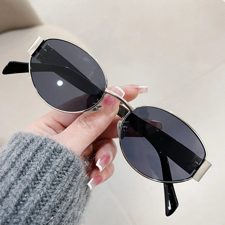 A hand with long, manicured nails holds a pair of vintage-inspired sunglasses featuring oval, black lenses and thin metal frames. The person wears a gray ribbed sweater, with a light, softly blurred background.