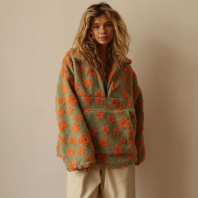 A woman with wavy blonde hair wears an autumn winter green flannel pullover with orange floral patterns and beige pants, standing against a plain light-colored background.
