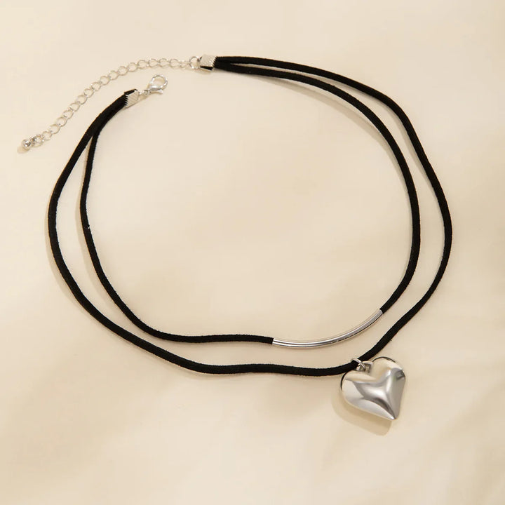 A black double cord velvet choker necklace featuring a shiny silver vintage heart pendant on one layer and a curved tube on the other, shown on a light beige background.