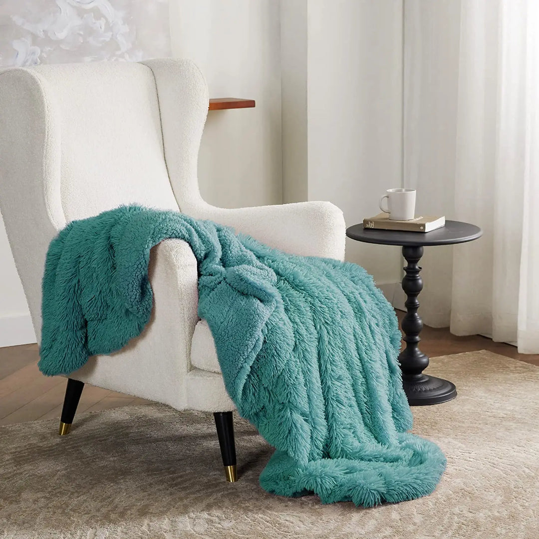 A white armchair with a sage green faux fur throw blanket draped over it sits next to a black round side table holding a white mug, in a cozy living room with light walls and a beige rug.