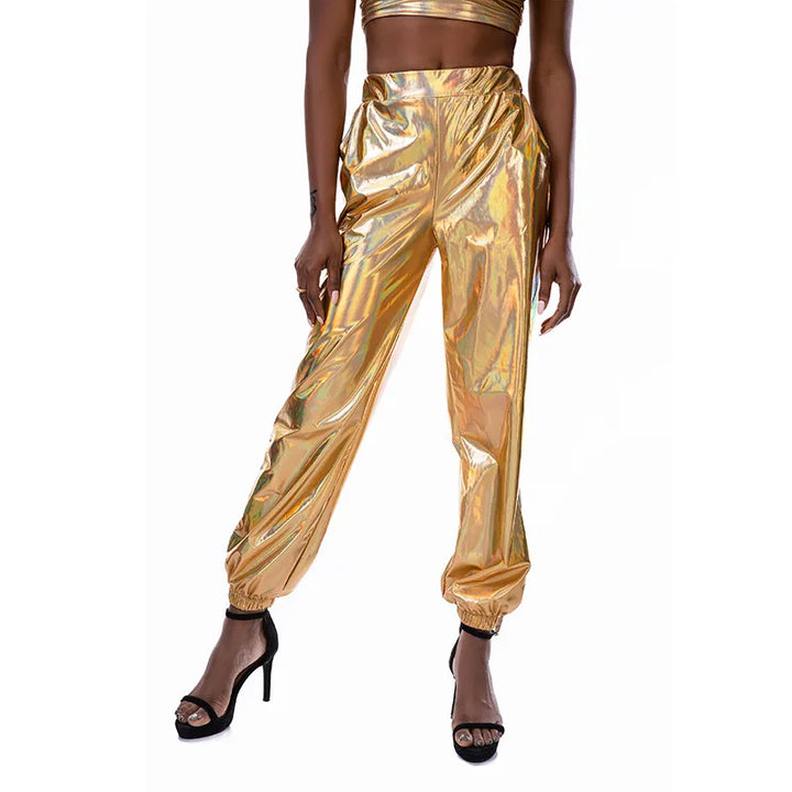 A person wearing shiny gold metallic jogger pants with an elastic high waist and loose fit, paired with black high-heeled sandals and a matching gold crop top partially visible. The background is plain white.