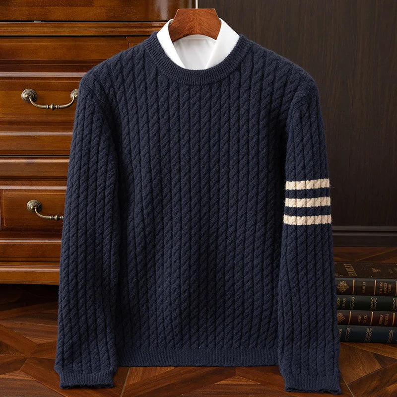 A navy blue cable knit sweater with ribbed cuffs and white stripes on one sleeve, displayed on a hanger over a collared shirt, placed in front of a wooden chest of drawers and stacked books.