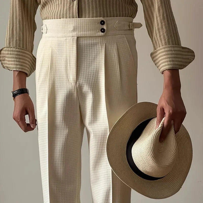 A person wearing a beige striped shirt and luxury tailored pants holds a straw hat with a black band in one hand. Their other hand shows a black wristwatch. The image is cropped at the shoulders.