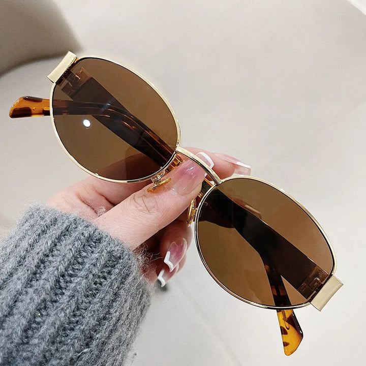 A hand with manicured nails holds a pair of vintage-inspired sunglasses—oval, gold-framed with brown tinted lenses and tortoiseshell arms—against a neutral background.