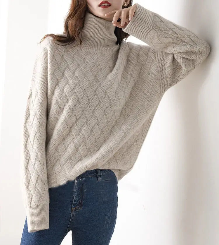 A person wearing an ultra soft, beige cable-knit turtleneck sweater and blue jeans, posing with one hand touching their chin against a white background.