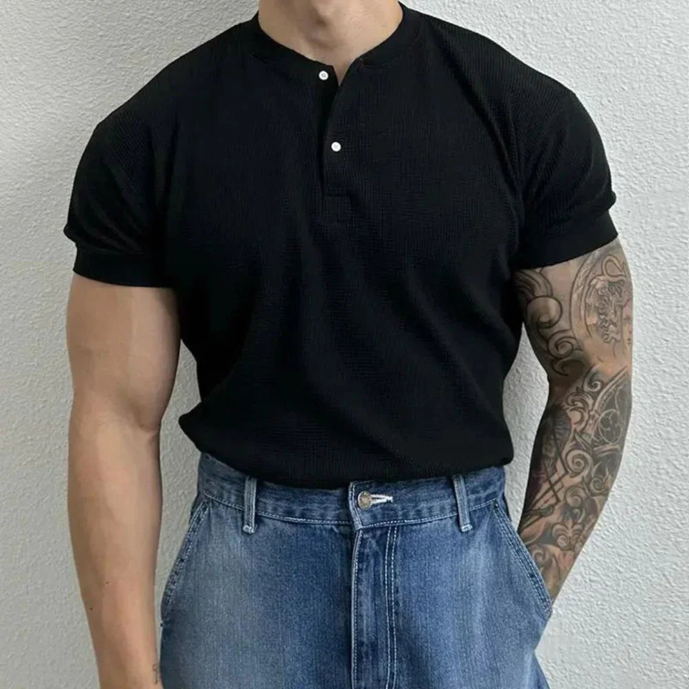 A person wearing a black, form-fitting shirt with a Henley neckline and blue jeans stands in front of a light-colored textured wall. One arm is tattooed with an intricate sleeve design. The person’s head is not visible.