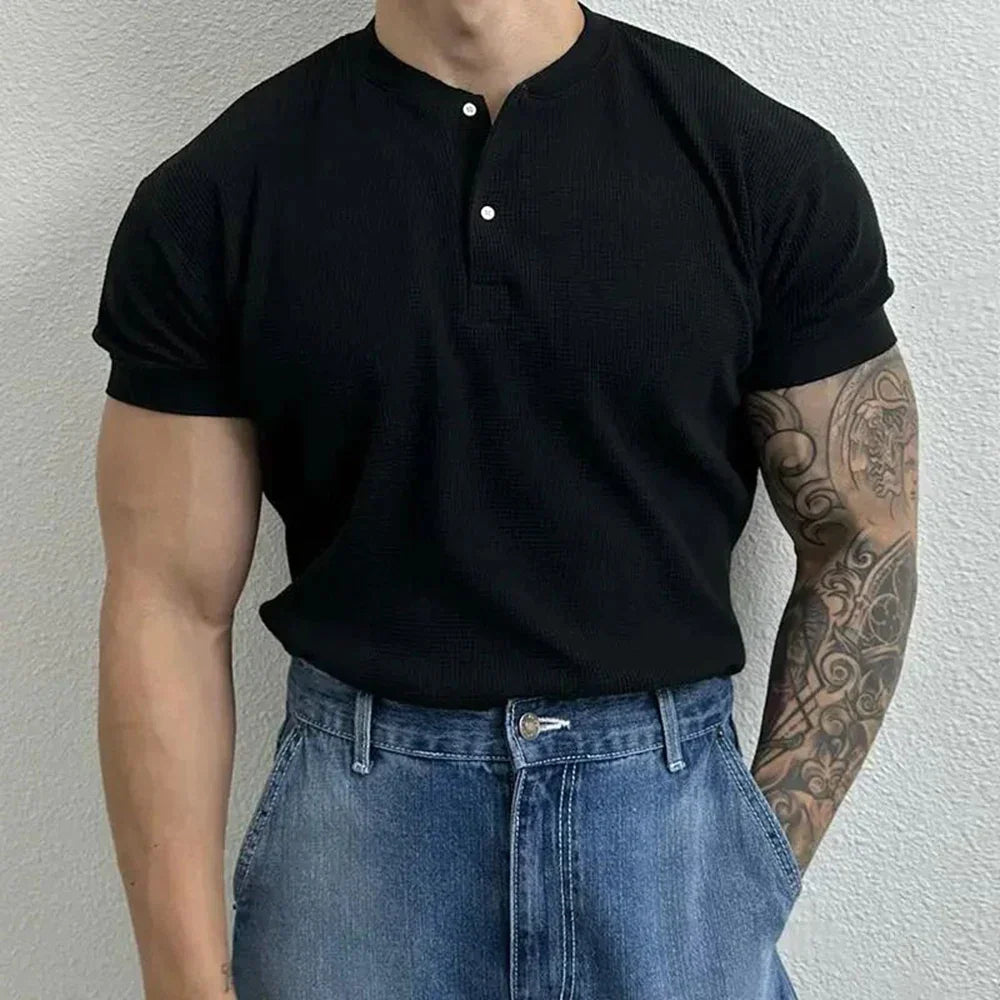 A person wearing a ribbed Henley short-sleeved black shirt and high-waisted blue jeans stands against a light-colored textured wall. Their right arm is tattooed with intricate designs. Their head is out of frame.