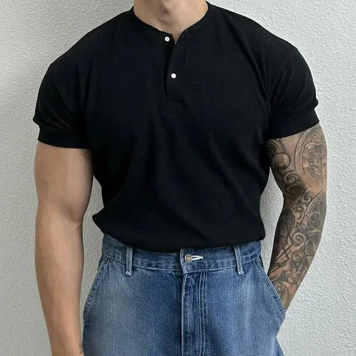A person wearing a ribbed Henley short-sleeved black shirt and high-waisted blue jeans stands against a light-colored textured wall. Their right arm is tattooed with intricate designs. Their head is out of frame.