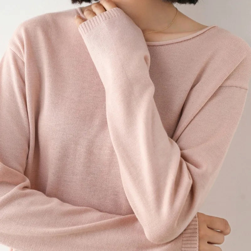 A person with short dark hair wears a light pink, long-sleeve cashmere blend sweater in a relaxed fit. The image is cropped at the shoulders, and their hand partially covers their mouth. The background is plain and light-colored.