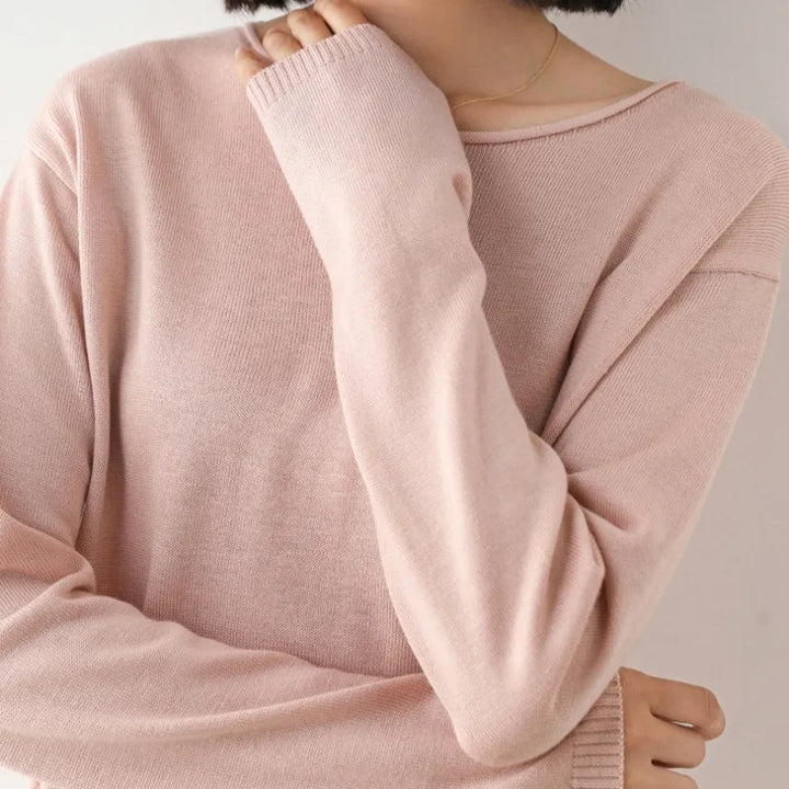 A person with short dark hair wears a light pink, long-sleeve cashmere blend sweater in a relaxed fit. The image is cropped at the shoulders, and their hand partially covers their mouth. The background is plain and light-colored.