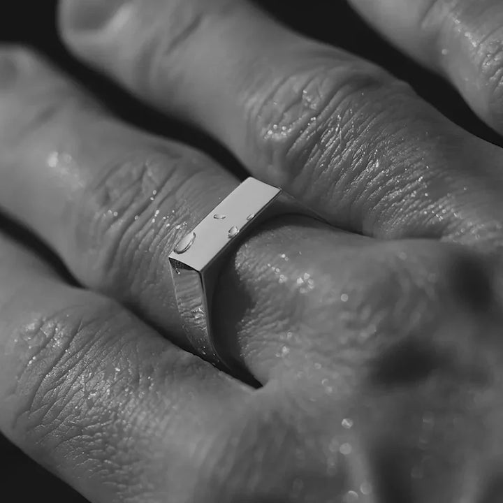 Close-up of a hand wearing a rectangular stainless steel ring, with small water droplets on the ring and skin, photographed in black and white.