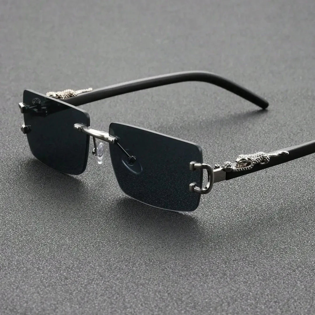 Rimless black sunglasses with rectangular lenses and decorative silver dragon details on the arms, featuring durable hinges, displayed on a textured dark gray surface.