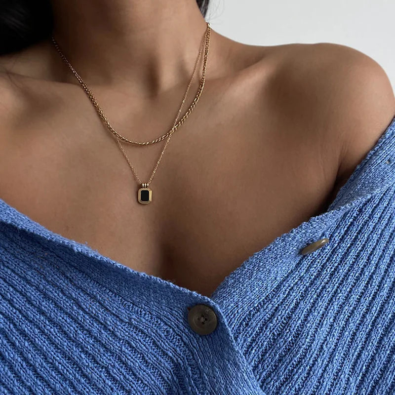 A person wearing a textured blue cardigan off the shoulders, showing two layered gold necklaces—one with a small square pendant—against bare skin and a neutral background.