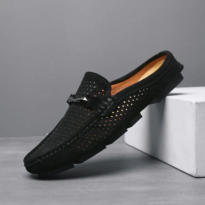 A black perforated slip-on loafer with a textured sole and decorative metal detail rests with its heel elevated on a white block against a gray background—perfect breathable loafers for effortless summer style.