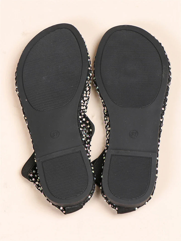 A pair of black flat sandals shown from the bottom, displaying textured rubber soles and rhinestone accents visible along the edges.