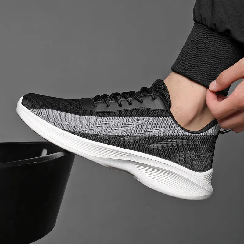 A person wearing a black and gray athletic sneaker with white sole, designed with breathable mesh for comfort, is shown in a close-up against a plain gray background. The foot is slightly lifted as if stepping onto a surface.