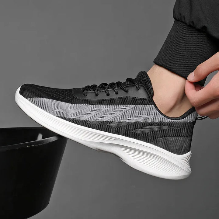 A person wearing a black and gray athletic sneaker with white sole, designed with breathable mesh for comfort, is shown in a close-up against a plain gray background. The foot is slightly lifted as if stepping onto a surface.