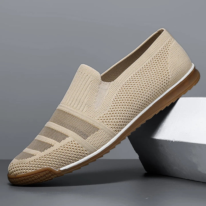 A beige slip-on men's casual loafer with a knitted mesh design, canvas lining, white midsole, and brown rubber outsole, displayed against a gray background and resting on a geometric block.