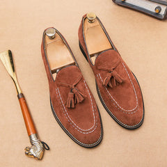 A pair of brown suede tassel loafers with cushioned footbed, white stitching, and wooden shoe trees are displayed. A shoehorn with a decorative handle rests beside them on a light brown surface.