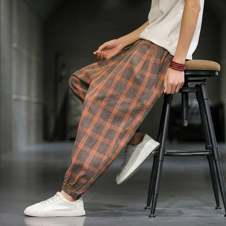 Person sitting sideways on a stool, wearing a white shirt, loose brown checkered pants with orange stripes inspired by Tokyo streetwear, white sneakers, and red beaded bracelets in a minimalist indoor setting.