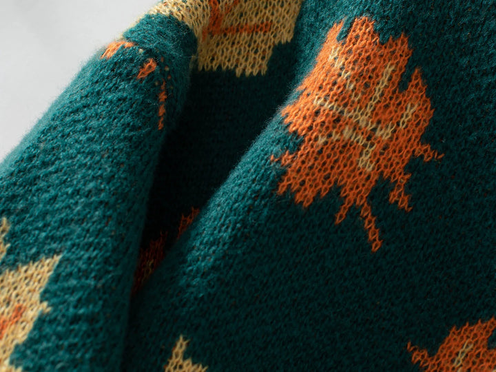 Close-up of a green soft knit material featuring an orange and yellow maple leaf pattern. The texture of the knitting and the detailed leaf designs are clearly visible on this comfortable sweater.