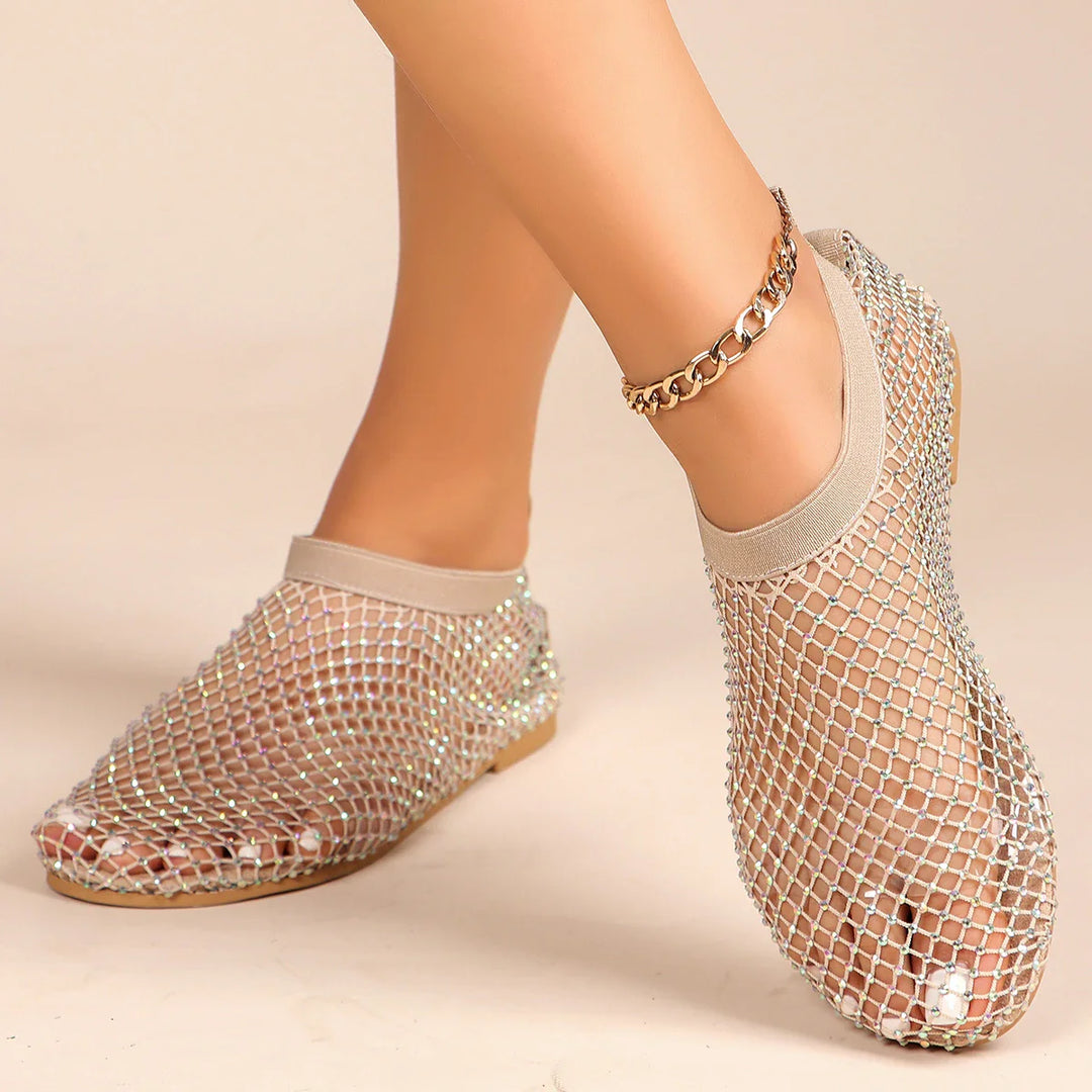 A person wearing mesh overlay shoes with rhinestone accents and a gold chain anklet stands on a neutral background.