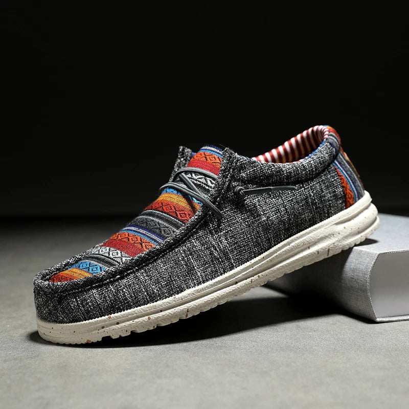 A casual slip-on shoe with a gray woven upper, colorful geometric patterns on the top, a white sole, and striped red-and-white inner lining, displayed on a gray surface with part of the sole resting on a block.