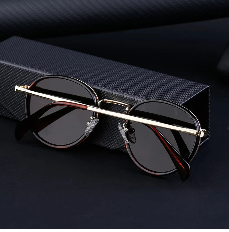 A pair of rounded sunglasses with dark, scratch resistant lenses and gold metal frames rest on a textured black case, set against a black background.