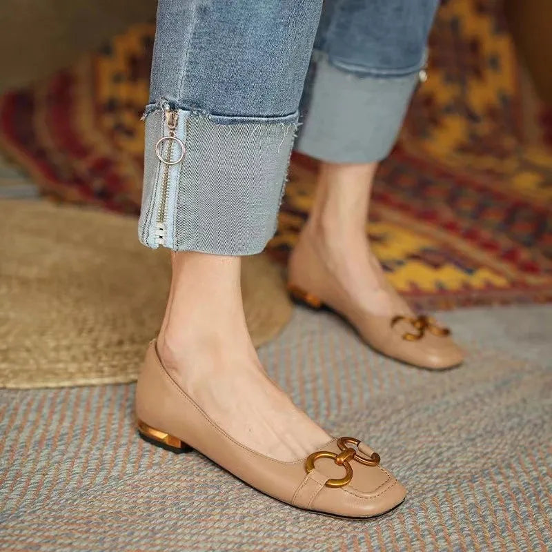 A person wearing light brown flats with metallic accents and gold decorative buckles, paired with cuffed blue jeans featuring zipper details, stands on a patterned rug.