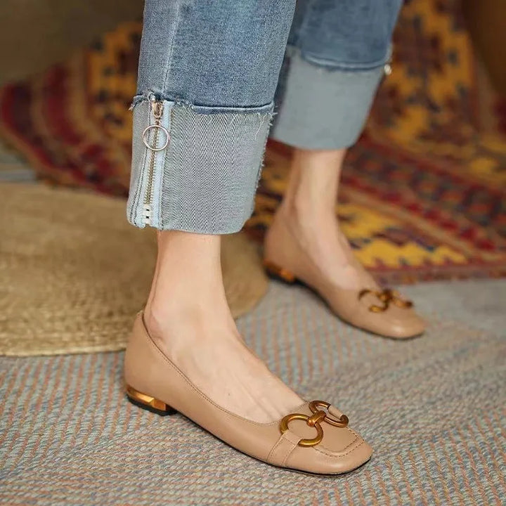 A person wearing light brown flats with metallic accents and gold decorative buckles, paired with cuffed blue jeans featuring zipper details, stands on a patterned rug.