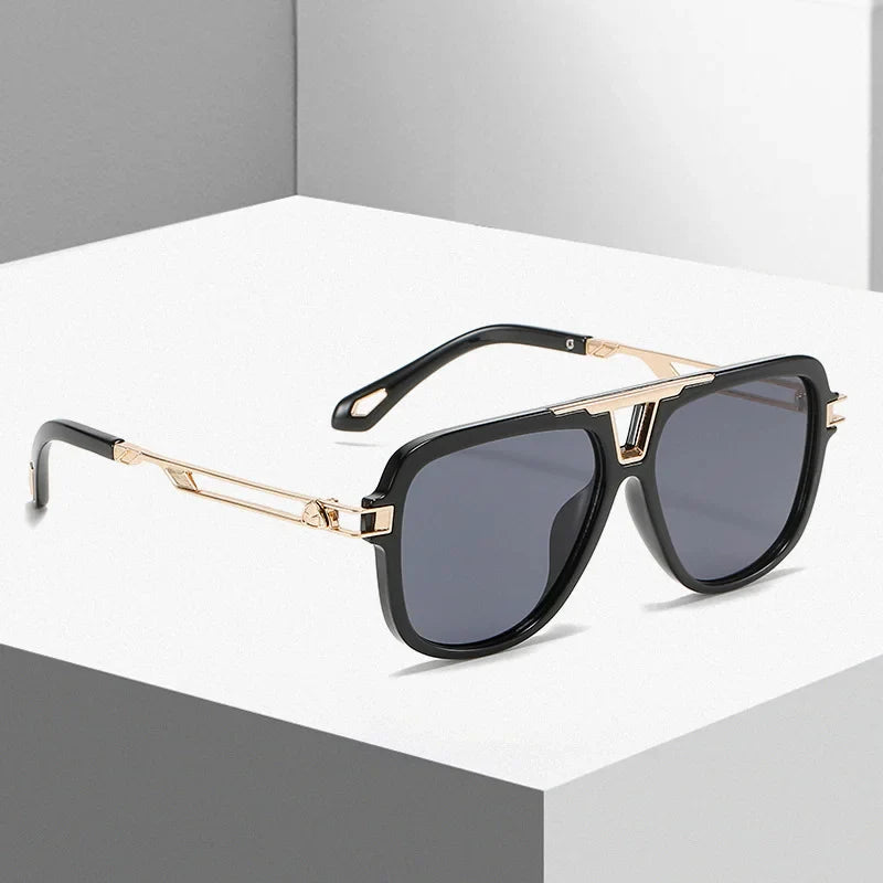 A pair of modern black luxury designer sunglasses with gold metal accents on the temples and a geometric frame, resting on a white and gray minimalist surface.