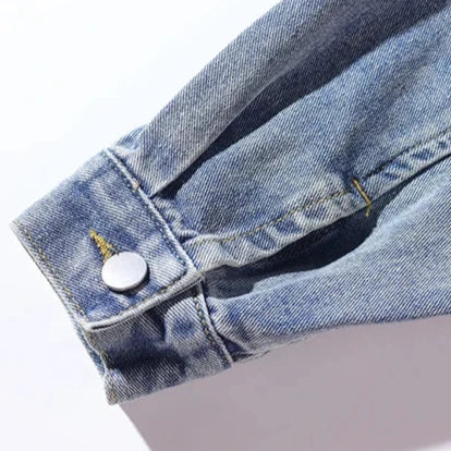 A close-up of the cuff of a light blue denim jacket sleeve, featuring a silver button and visible yellow stitching, highlights its oversized fit and subtle retro style.