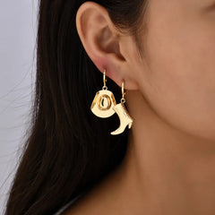 Close-up of a woman’s ear adorned with mismatched gold earrings—a cowboy hat and a boot—showcasing stylish western jewelry, with long dark hair partially visible.