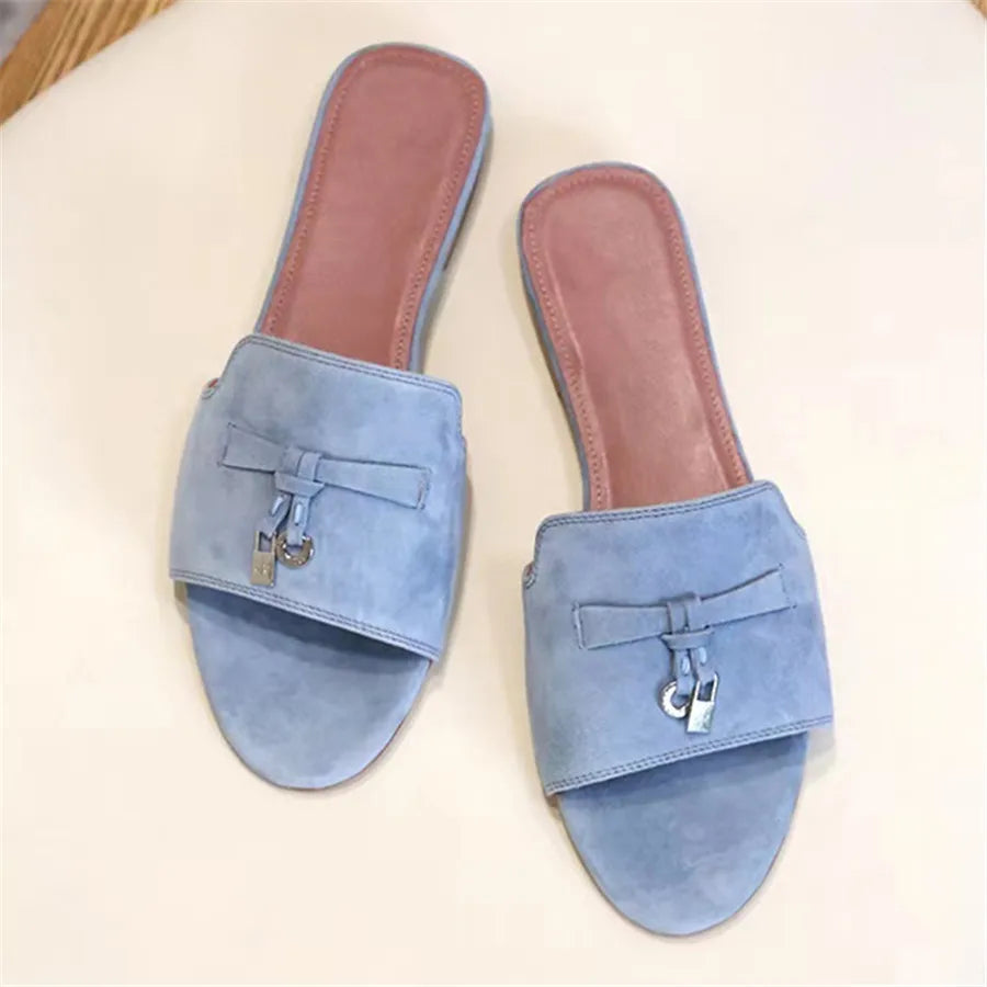 A pair of light blue suede finish slide sandals with a closed toe strap, cushioned pink insoles, and decorative silver buckle details on each sandal, placed on a light surface.