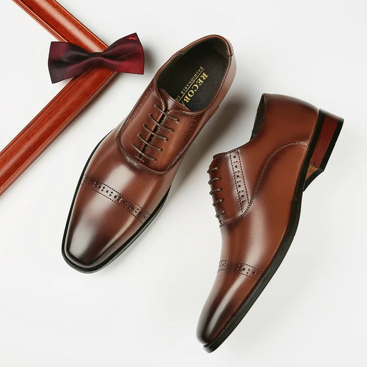 A pair of polished brown Italian leather dress shoes with laces is displayed on a white surface next to a dark red bow tie and the corner of a wooden frame.