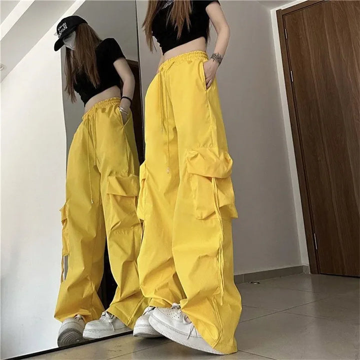 A person wearing a black crop top, black cap, bright yellow high-waist cargo pants with adjustable drawstring waist and cargo pockets, and white sneakers stands in front of a mirror in a minimal indoor setting. The reflection shows the full outfit.