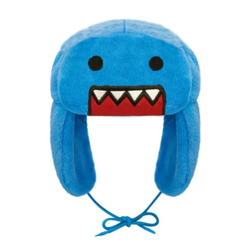 A blue plush hat with adjustable ear flaps, featuring a cartoon face with black eyes and a red mouth with white triangular teeth. The hat has a string tie at the bottom for cozy winter styling.