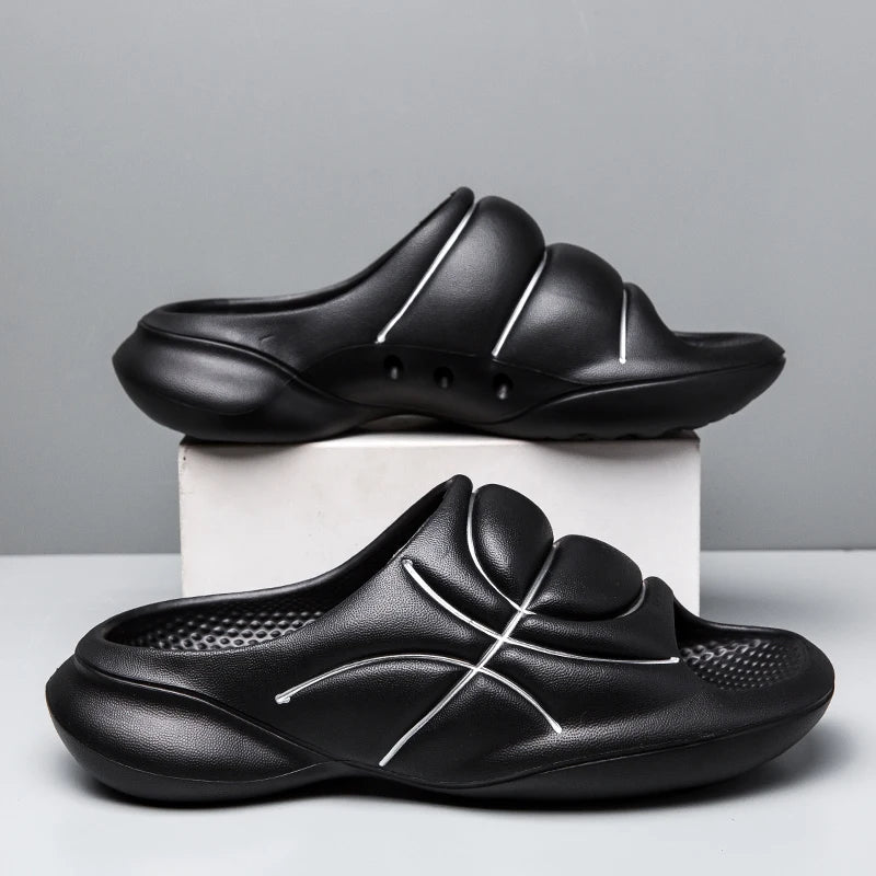 A pair of black basketball slip ons with thick, cushioned soles and white curved line designs are displayed, one on a white box and the other on the surface, against a plain gray background.