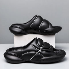 A pair of black basketball slip ons with thick, cushioned soles and white curved line designs are displayed, one on a white box and the other on the surface, against a plain gray background.