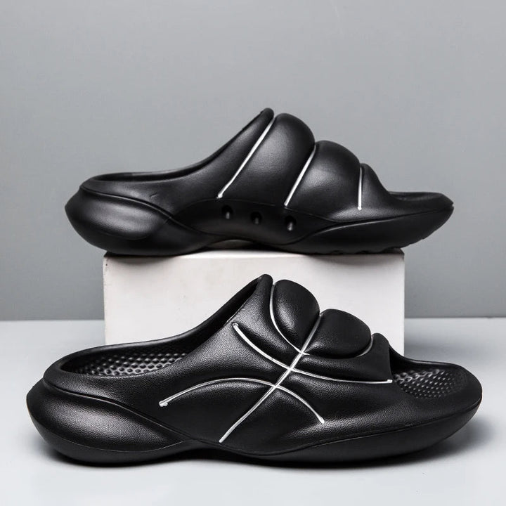 A pair of black basketball slip ons with thick, cushioned soles and white curved line designs are displayed, one on a white box and the other on the surface, against a plain gray background.