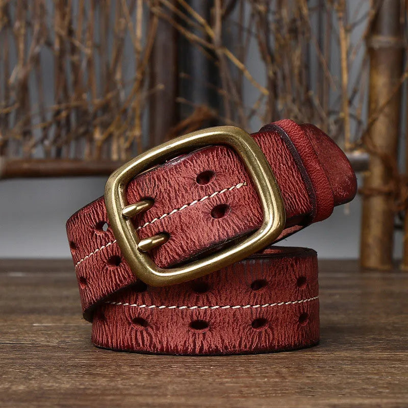 A genuine leather belt with a textured surface, decorative stitching, oval-shaped holes, and a large brass buckle showcases heritage craftsmanship, displayed coiled on a wooden surface with branches in the background.