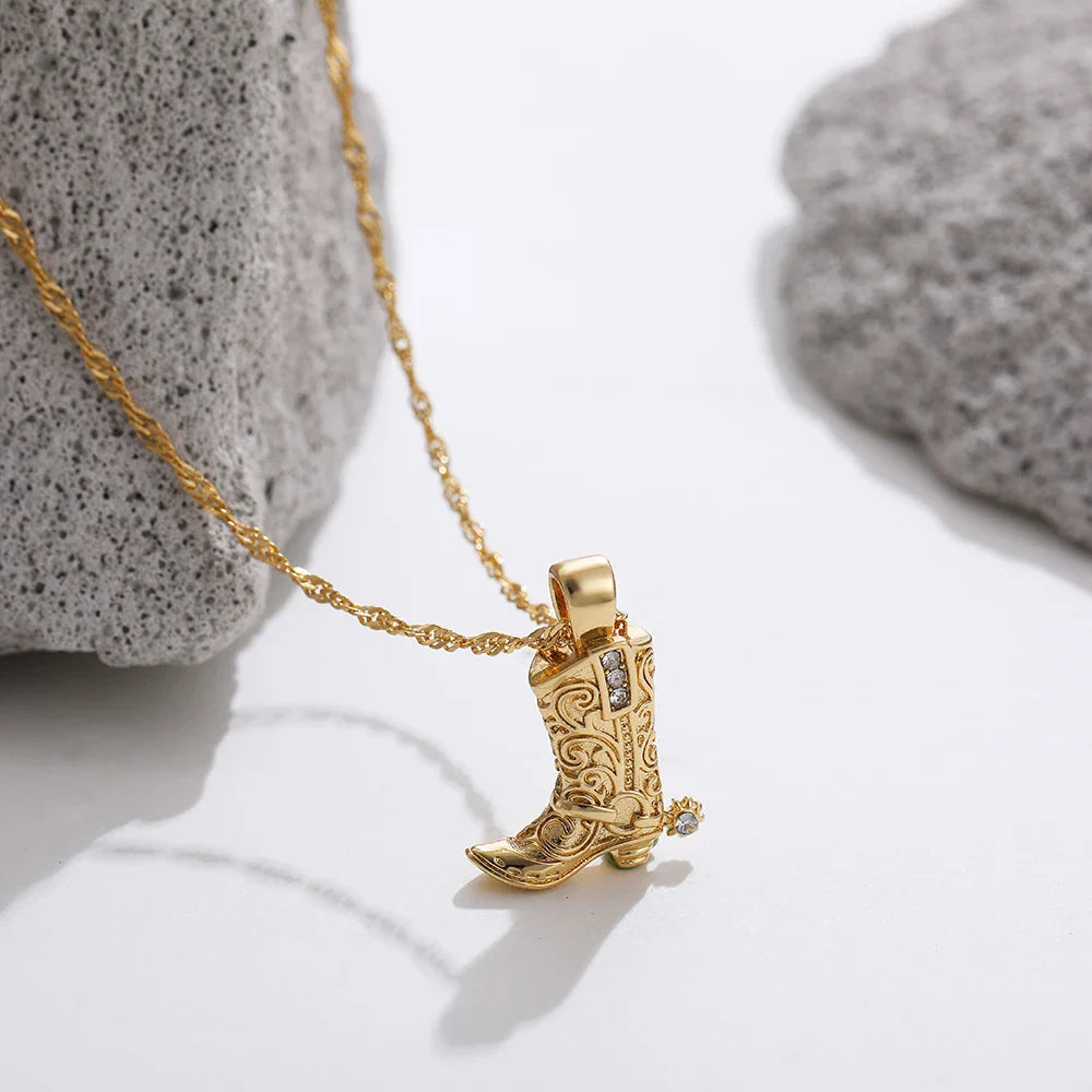 A gold cowgirl necklace with a detailed cowboy boot pendant, featuring intricate designs, is displayed on a white surface near two rough gray stones.