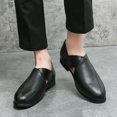A person wearing genuine leather loafers and black pants stands on a gray concrete floor with a stone-textured wall in the background, highlighting the shoes durable outsole.