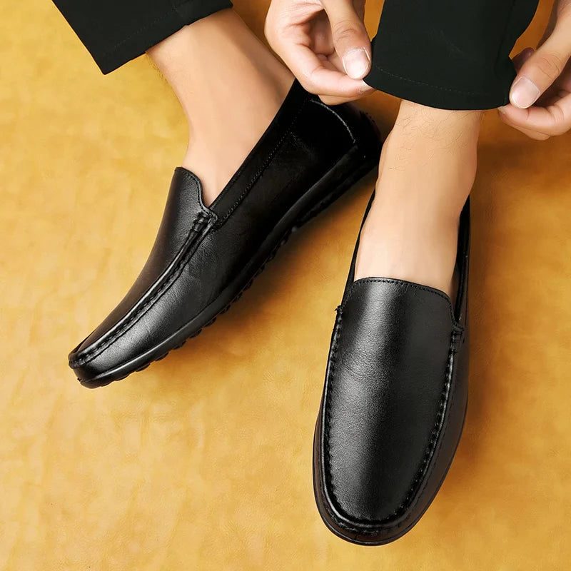 A person wearing black leather loafers with a cushioned footbed sits on a yellow-brown background, adjusting one shoe. The loafers have a sleek design and visible stitching along the edges.