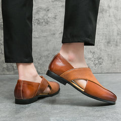 A person wearing low profile loafers with textured brown genuine leather straps and durable outsoles, paired with black pants, stands on a gray textured floor against a gray wall.
