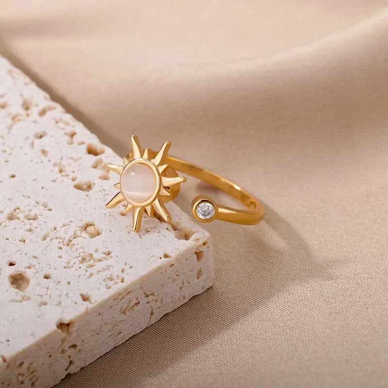 A sun ring with a gold sun-shaped design features a central round stone and a small clear gem on the adjustable gold ring band, displayed on a light textured surface against beige fabric.
