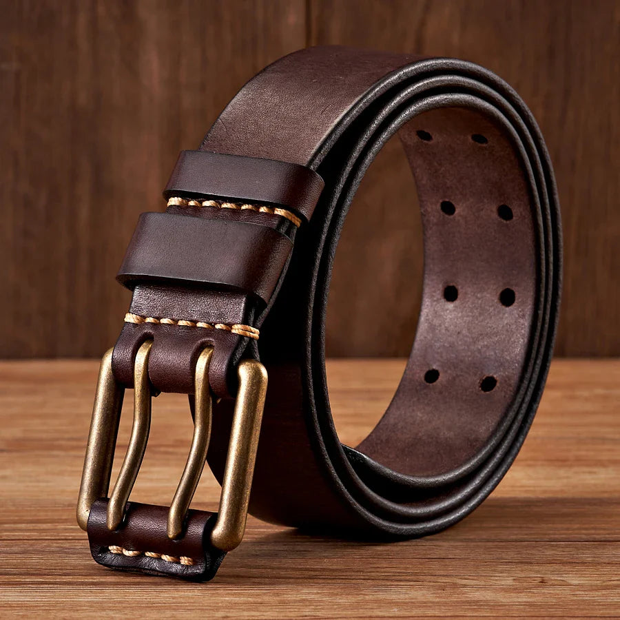 A rolled-up dark brown cowhide leather belt with a double copper buckle and contrast stitching, displayed on a wooden surface with a wood-paneled background.